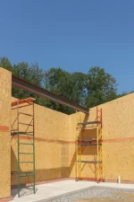 Цех «Евгений» 400 м², Шанырак (Алматы), 2026: стальная балка уложена по верху стен из СИП-панелей во внутреннем углу цеха, у стен подняты две передвижные вышки-туры