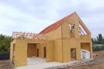 Дом «Игорь» 200 м², Талгар, 2026: скат крыши над двухэтажным объёмом зашит панелями по стропилам, над пристроем стропила пока открыты, во фронтоне виден проём мансардного окна с раскосами