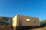 Дом «Пётр» 54 м², Каскелен, 2026: коробка дома собрана — наружные стены из СИП-панелей выведены на полную высоту, видны дверной проём и окна, цоколь покрыт битумной гидроизоляцией, на заднем плане предгорья