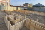 Дом «Сатурин» 165 м², Алматы, 2026: опалубка вблизи — угол ленты с арматурным каркасом и подкосами, за оградой соседние дома