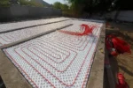 Дом «Сатурин» 165 м², Алматы, 2026: вид вдоль плиты — маты с уложенной трубой тёплого пола, у борта фундамента бухты красной трубы под разводку