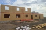 Общежитие «Доссан» 550 м², Астана, 2026: длинный фасад с проёмами под окна и двери, на перекрытии сложены пачки СИП-панелей, рядом штабель бруса