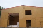 Цех «Евгений» 400 м², Шанырак (Алматы), 2026: торцевая стена цеха выведена до фронтона, по верху уложены обвязочные брусья, над воротным проёмом видна стропильная система с настилом кровли, оставлены проёмы под ворота и окна