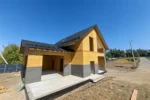 Дом «Игорь» 200 м², Талгар, 2026: кровля из металлочерепицы смонтирована на обоих объёмах дома, нижняя часть стен закрыта ветрозащитной мембраной, залита плита террасы и ступени крыльца
