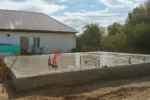 Дом «Надежда» 100 м², Павлодар, 2026: вид со стороны дома — стяжка основания залита, из неё выведены выпуски канализации и водопровода, пучок красных труб под разводку