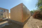 Дом «Сатурин» 165 м², Алматы, 2026: угол дома — две наружные стены из СИП-панелей сошлись на угловой стойке, по верху лент видна гидроизоляция цоколя