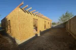 Дом «Сатурин» 165 м², Алматы, 2026: вид с угла — стены собраны по всему периметру, проёмы под окна и двери оставлены, балки перекрытия уложены с равным шагом