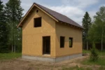 Баня «Н-90», 90 м², 1,5 этажа, в черновой отделке: коробка из СИП-панелей OSB-3, вальмовая кровля из металлочерепицы, проёмы без окон и дверей, в проёмах видны сосновые балки перекрытия