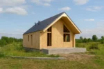 Дом «Б-104», 104 м², 1.5 этажа, в черновой отделке: коробка из СИП-панелей OSB-3, двускатная кровля из металлочерепицы, проёмы без окон и дверей, в проёмах видны сосновые балки перекрытия