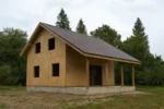 Дом «Б-131В», 131 м², 1.5 этажа, в черновой отделке: коробка из СИП-панелей OSB-3, двускатная кровля из металлочерепицы, проёмы без окон и дверей, в проёмах видны сосновые балки перекрытия
