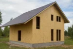 Дом «Б-160Б», 160 м², 1.5 этажа, в черновой отделке: коробка из СИП-панелей OSB-3, двускатная кровля из металлочерепицы, проёмы без окон и дверей, в проёмах видны сосновые балки перекрытия
