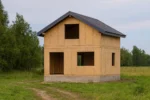 Дом «Д-102Б», 102 м², 1.5 этажа, в черновой отделке: коробка из СИП-панелей OSB-3, двускатная кровля из металлочерепицы, проёмы без окон и дверей, в проёмах видны сосновые балки перекрытия