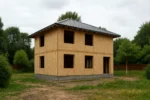 Дом «Ё-152», 152 м², 2 этажа, в черновой отделке: коробка из СИП-панелей OSB-3, вальмовая кровля из металлочерепицы, проёмы без окон и дверей, в проёмах видны сосновые балки перекрытия