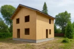 Дом «Е-164», 164 м², 2 этажа, в черновой отделке: коробка из СИП-панелей OSB-3, двускатная кровля из металлочерепицы, проёмы без окон и дверей, в проёмах видны сосновые балки перекрытия