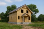 Дом «Г-168Б», 168 м², 1.5 этажа, в черновой отделке: коробка из СИП-панелей OSB-3, двускатная кровля из металлочерепицы, проёмы без окон и дверей, в проёмах видны сосновые балки перекрытия