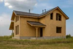 Дом «Г-182», 182 м², 1.5 этажа, в черновой отделке: коробка из СИП-панелей OSB-3, двускатная кровля из металлочерепицы, проёмы без окон и дверей, в проёмах видны сосновые балки перекрытия