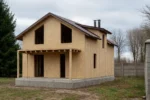 Дом «К-164Б», 164 м², 1.5 этажа, в черновой отделке: коробка из СИП-панелей OSB-3, двускатная кровля из металлочерепицы, проёмы без окон и дверей, в проёмах видны сосновые балки перекрытия