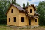 Дом «К-171В», 171 м², 1.5 этажа, в черновой отделке: коробка из СИП-панелей OSB-3, двускатная кровля из металлочерепицы, проёмы без окон и дверей, в проёмах видны сосновые балки перекрытия