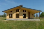 Дом «К-229Б», 229 м², 1.5 этажа, в черновой отделке: коробка из СИП-панелей OSB-3, двускатная кровля из металлочерепицы, проёмы без окон и дверей, в проёмах видны сосновые балки перекрытия