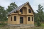 Дом «Л-110», 110 м², 1.5 этажа, в черновой отделке: коробка из СИП-панелей OSB-3, двускатная кровля из металлочерепицы, проёмы без окон и дверей, в проёмах видны сосновые балки перекрытия