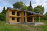 Дом «М-135В», 135 м², 1.5 этажа, в черновой отделке: коробка из СИП-панелей OSB-3, двускатная кровля из металлочерепицы, проёмы без окон и дверей, в проёмах видны сосновые балки перекрытия