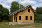 Дом «Н-84В», 84 м², 1.5 этажа, в черновой отделке: коробка из СИП-панелей OSB-3, двускатная кровля из металлочерепицы, проёмы без окон и дверей, в проёмах видны сосновые балки перекрытия