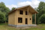 Дом «О-112Б», 112 м², 1.5 этажа, в черновой отделке: коробка из СИП-панелей OSB-3, двускатная кровля из металлочерепицы, проёмы без окон и дверей, в проёмах видны сосновые балки перекрытия