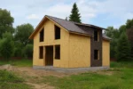 Дом «Р-150Б», 150 м², 1.5 этажа, в черновой отделке: коробка из СИП-панелей OSB-3, двускатная кровля из металлочерепицы, проёмы без окон и дверей, в проёмах видны сосновые балки перекрытия