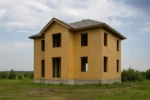 Дом «Р-157», 157 м², 2 этажа, в черновой отделке: коробка из СИП-панелей OSB-3, вальмовая кровля из металлочерепицы, проёмы без окон и дверей, в проёмах видны сосновые балки перекрытия