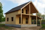 Дом «С-140Б», 140 м², 1.5 этажа, в черновой отделке: коробка из СИП-панелей OSB-3, двускатная кровля из металлочерепицы, проёмы без окон и дверей, в проёмах видны сосновые балки перекрытия