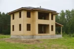 Дом «Т-112В», 112 м², 2 этажа, в черновой отделке: коробка из СИП-панелей OSB-3, вальмовая кровля из металлочерепицы, проёмы без окон и дверей, в проёмах видны сосновые балки перекрытия