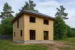 Дом «Т-180Б», 180 м², 2 этажа, в черновой отделке: коробка из СИП-панелей OSB-3, вальмовая кровля из металлочерепицы, проёмы без окон и дверей, в проёмах видны сосновые балки перекрытия