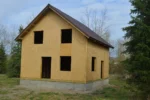 Дом «В-107Б», 107 м², 1.5 этажа, в черновой отделке: коробка из СИП-панелей OSB-3, двускатная кровля из металлочерепицы, проёмы без окон и дверей, в проёмах видны сосновые балки перекрытия
