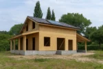 Дом «В-181Б», 181 м², 1.5 этажа, в черновой отделке: коробка из СИП-панелей OSB-3, двускатная кровля из металлочерепицы, проёмы без окон и дверей, в проёмах видны сосновые балки перекрытия
