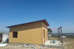 Дом «Айни» 50 м², Бирлик, 2026: коробка из СИП-панелей под односкатной кровлей из металлочерепицы, свесы с открытыми стропилами, справа — стальной каркас навеса
