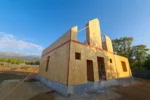 Дом «Игорь» 200 м², Талгар, 2026: коробка первого этажа из СИП-панелей собрана, поверх стен уложена обвязка, панели второго этажа подняты и раскреплены подкосами, на заднем плане горы