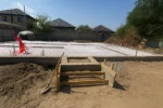 Дом «Сатурин» 165 м², Алматы, 2026: вид со стороны входа — тёплый пол разведён по всей плите, выпуски труб связаны в пучок, у фундамента собрана опалубка крыльца