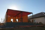 Дом «Надежда» 100 м², Павлодар, 2026: над собранными стенами смонтирован стальной каркас из колонн и балок, поверх него уложены деревянные балки перекрытия, вид против солнца
